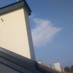 Nowy blaszany dach z kominem wykończonym obr&oacute;bką blacharską, na tle błękitnego nieba z lekkimi obłokami.