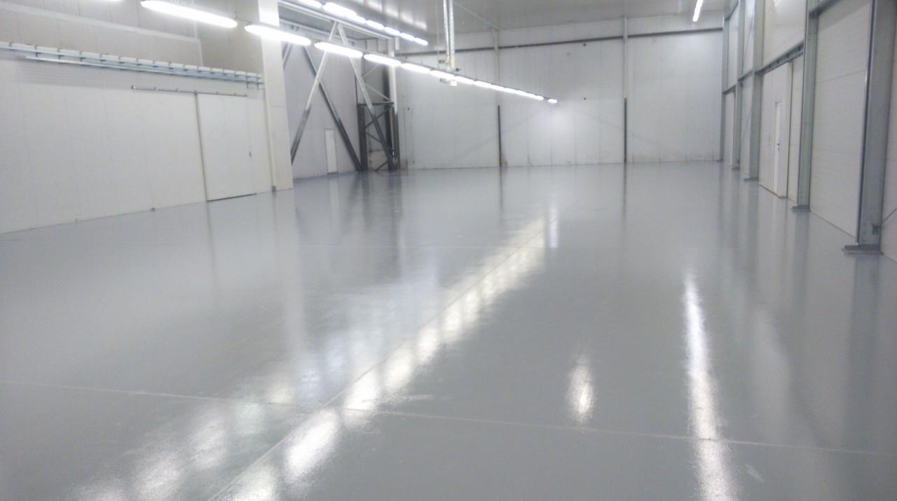 Błyszcząca, szara posadzka przemysłowa w hali magazynowej z białymi ścianami z paneli i oświetleniem ledowym. Widoczne metalowe słupy i drzwi przesuwne.
