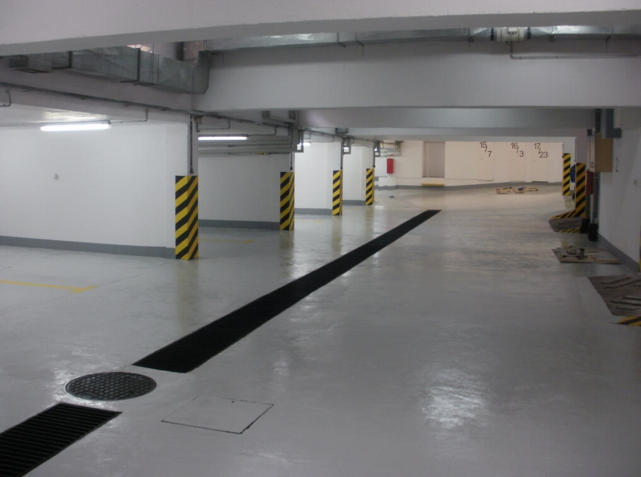 Jasnoszara posadzka garażu po malowaniu z czarnymi kratkami odpływowymi i słupami w pasy żółto-czarne. Świeżo pomalowane ściany i sufit.