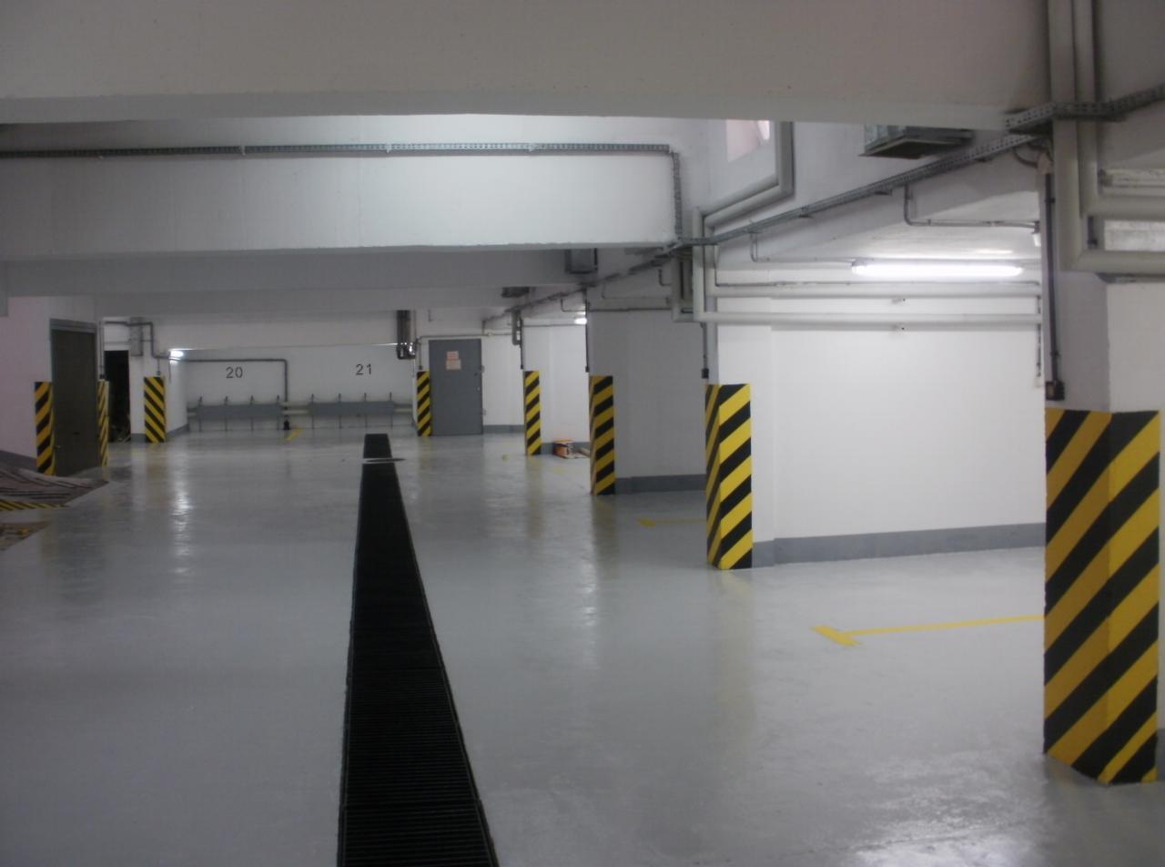 Szara posadzka w garażu podziemnym z czarno-żółtymi słupami i odpływem liniowym. Świeżo pomalowane ściany i podłoga, widoczne numery miejsc parkingowych.