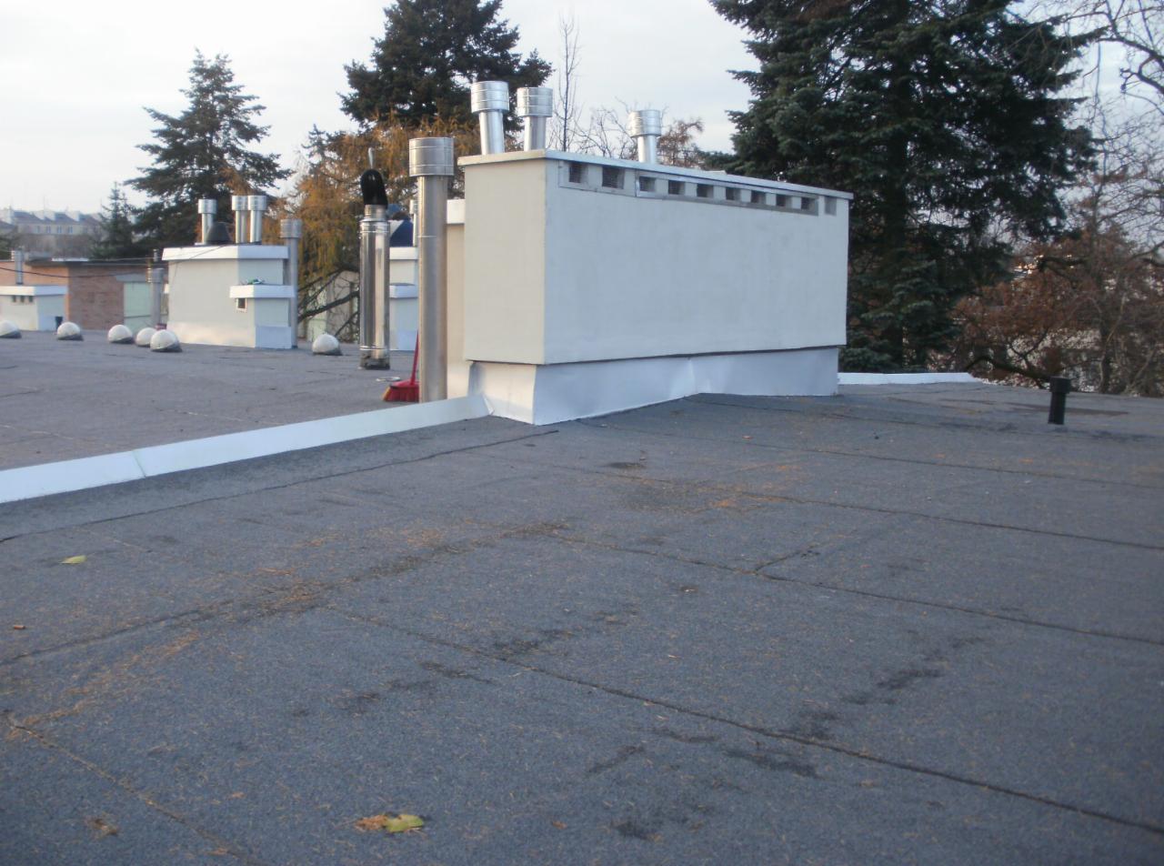 Widok na płaski dach pokryty papą, z kominami i elementami wentylacyjnymi. Widoczne obróbki blacharskie i uszczelnienia. Ujęcie z bliska, w tle drzewa i budynki.