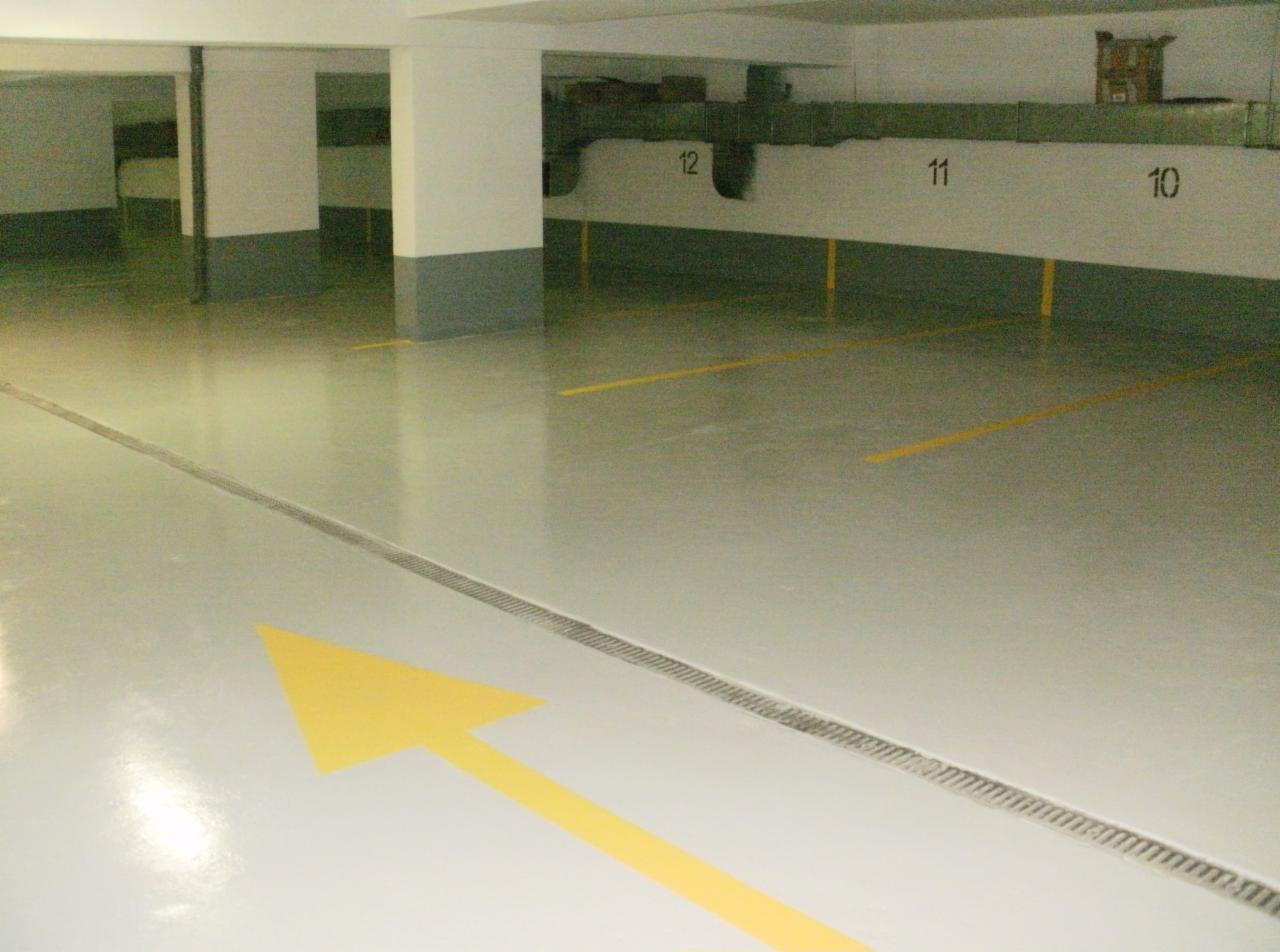 Jasne, lśniące posadzki w garażu podziemnym z żółtymi oznaczeniami miejsc parkingowych i strzałką kierunkową. Widoczne numery miejsc i odpływ liniowy.