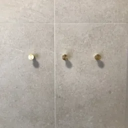 Jasnoszare płytki imitujące kamień z trzema złotymi, okrągłymi wieszakami na ręczniki, zamontowanymi na ścianie.