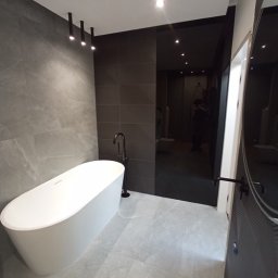 REMONTUJ Z NAMI - Nowoczesna łazienka w odcieniach szarości z wolnostojącą białą wanną, czarną armaturą i podłużnymi lampami sufitowymi, uzupełniona o ciemną ścianę z geometrycznym wzorem i lustrzaną taflę.