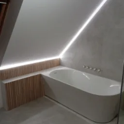 Nowoczesna łazienka na poddaszu z wolnostojącą wanną, podświetleniem LED wzdłuż skosu sufitu i drewnianą zabudową wanny.