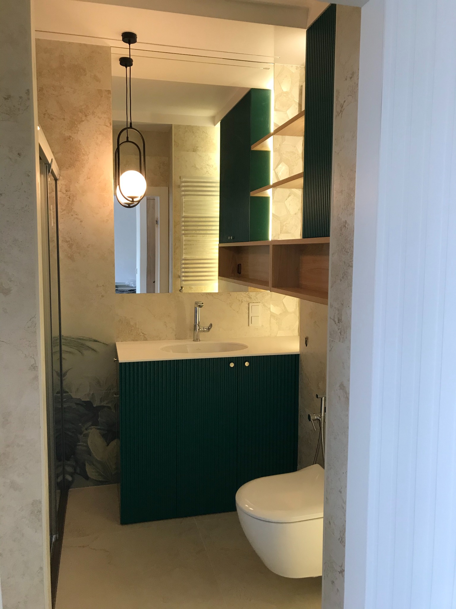 Nowoczesna łazienka z umywalką na szafce w kolorze butelkowej zieleni, wiszącą toaletą i dekoracyjną lampą wiszącą. Na ścianach marmurkowe płytki i akcent roślinny w kabinie prysznicowej.