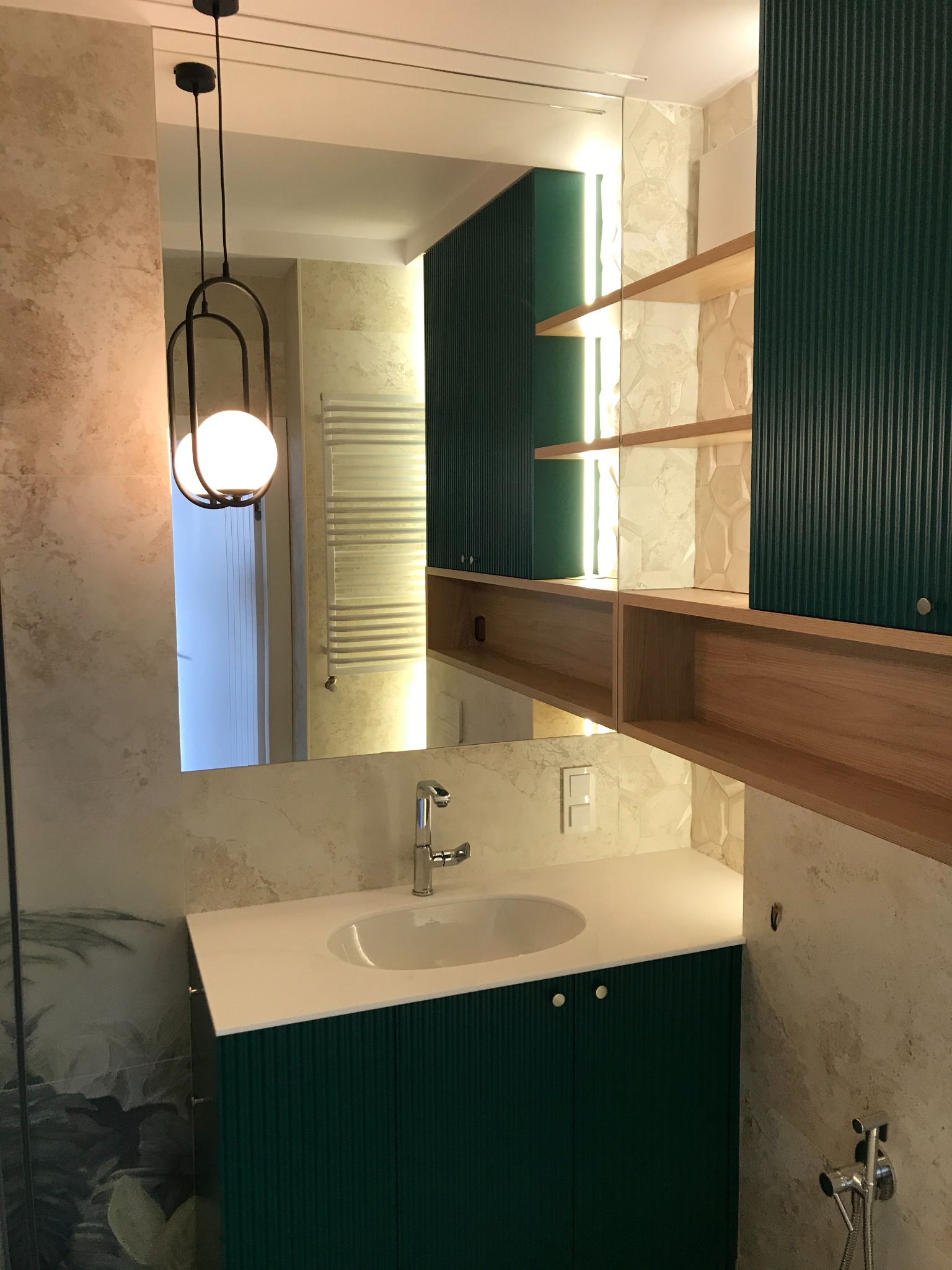Nowoczesna łazienka z umywalką nablatową na szafce w kolorze butelkowej zieleni, lustrem na ścianie w beżowych odcieniach, wiszącą lampą z kulistym kloszem i drewnianymi półkami z podświetleniem LED.