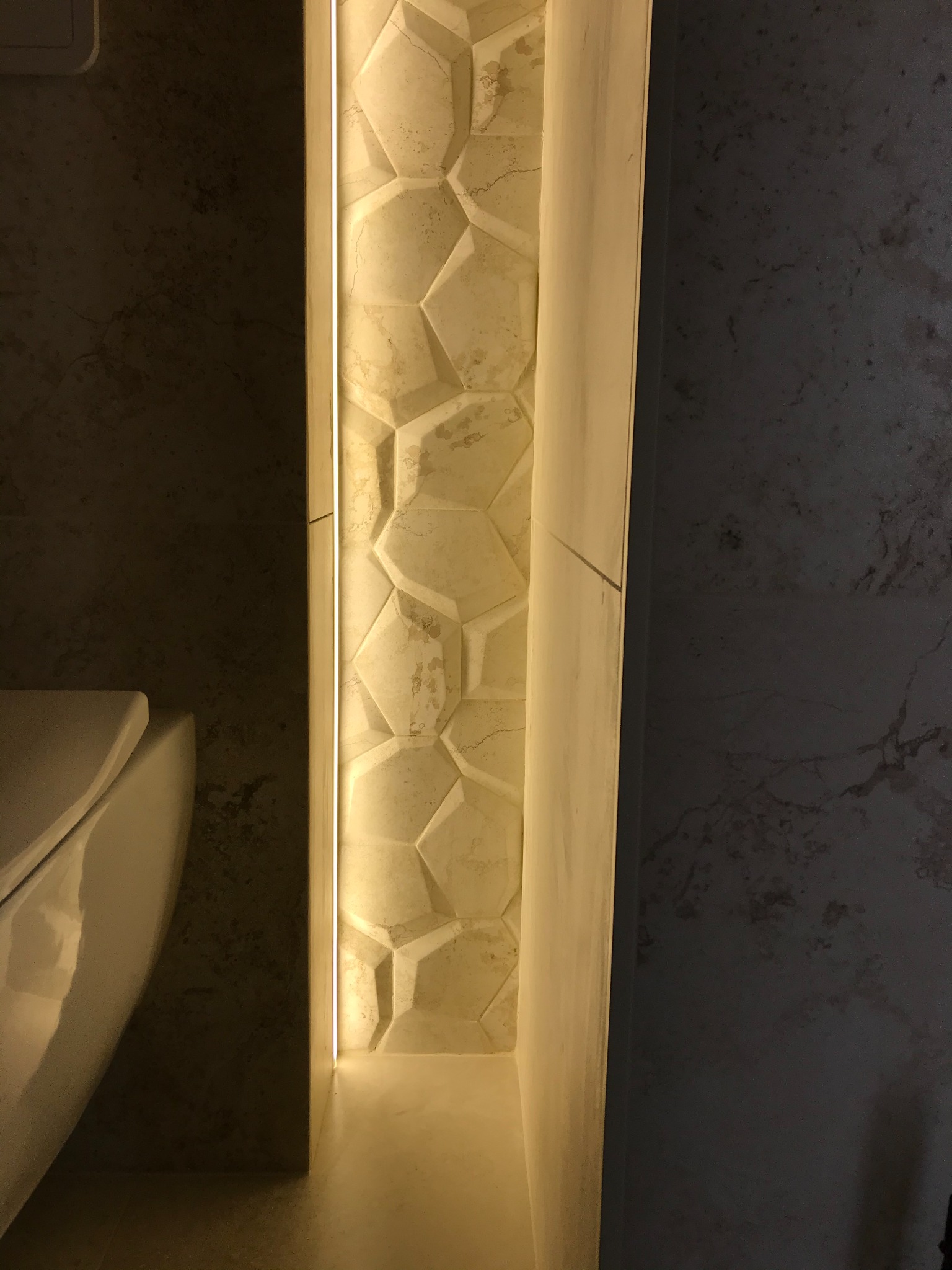 Narożnik łazienki z toaletą, dekoracyjną ścianą z heksagonalnych płytek podświetlonych ledowym paskiem i ścianą w ciemnym odcieniu lastryko.