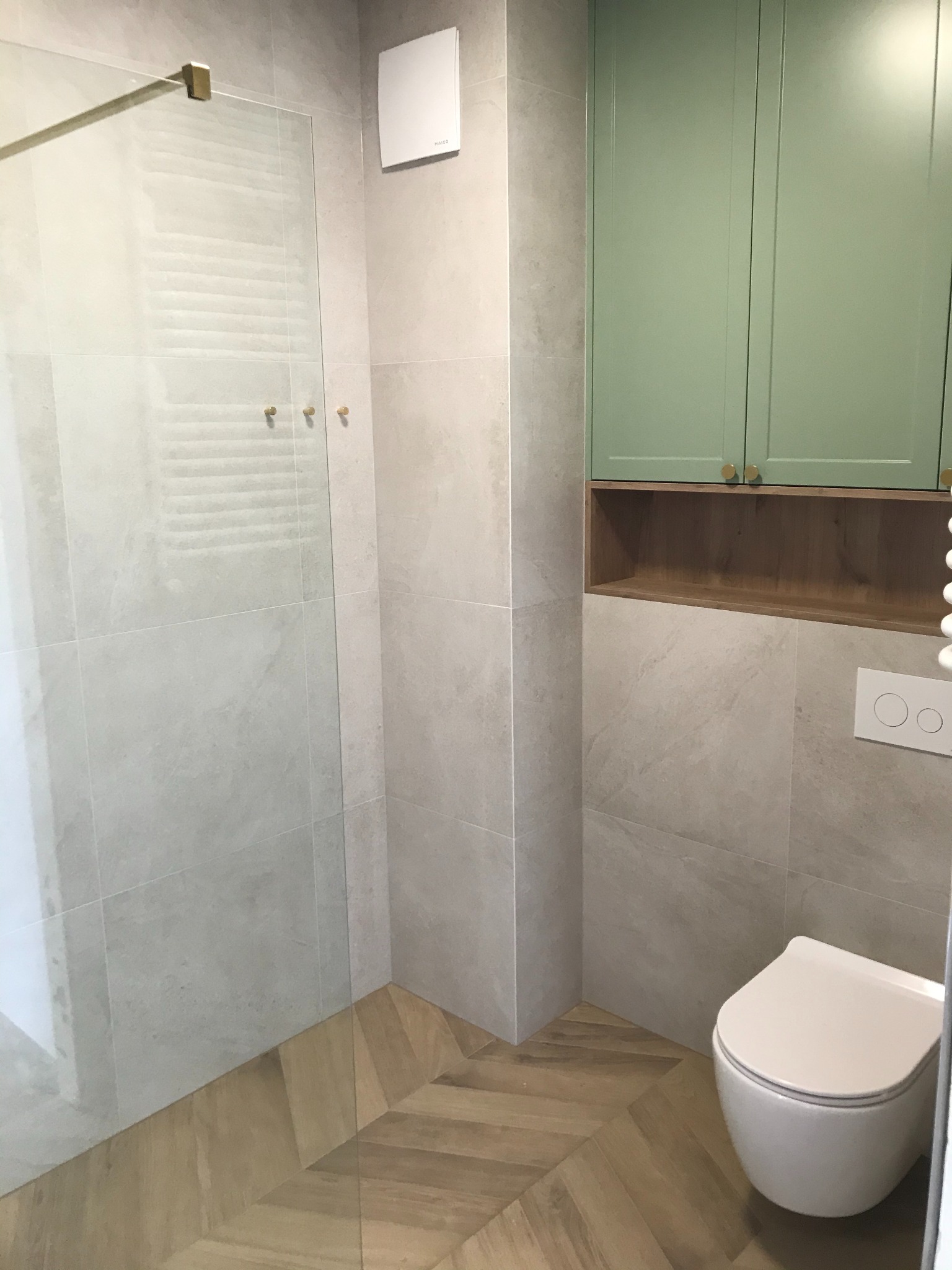 Nowoczesna łazienka z kabiną prysznicową ze złotymi detalami, płytkami imitującymi kamień, drewnianą podłogą w jodełkę i szafką w kolorze pistacjowym.