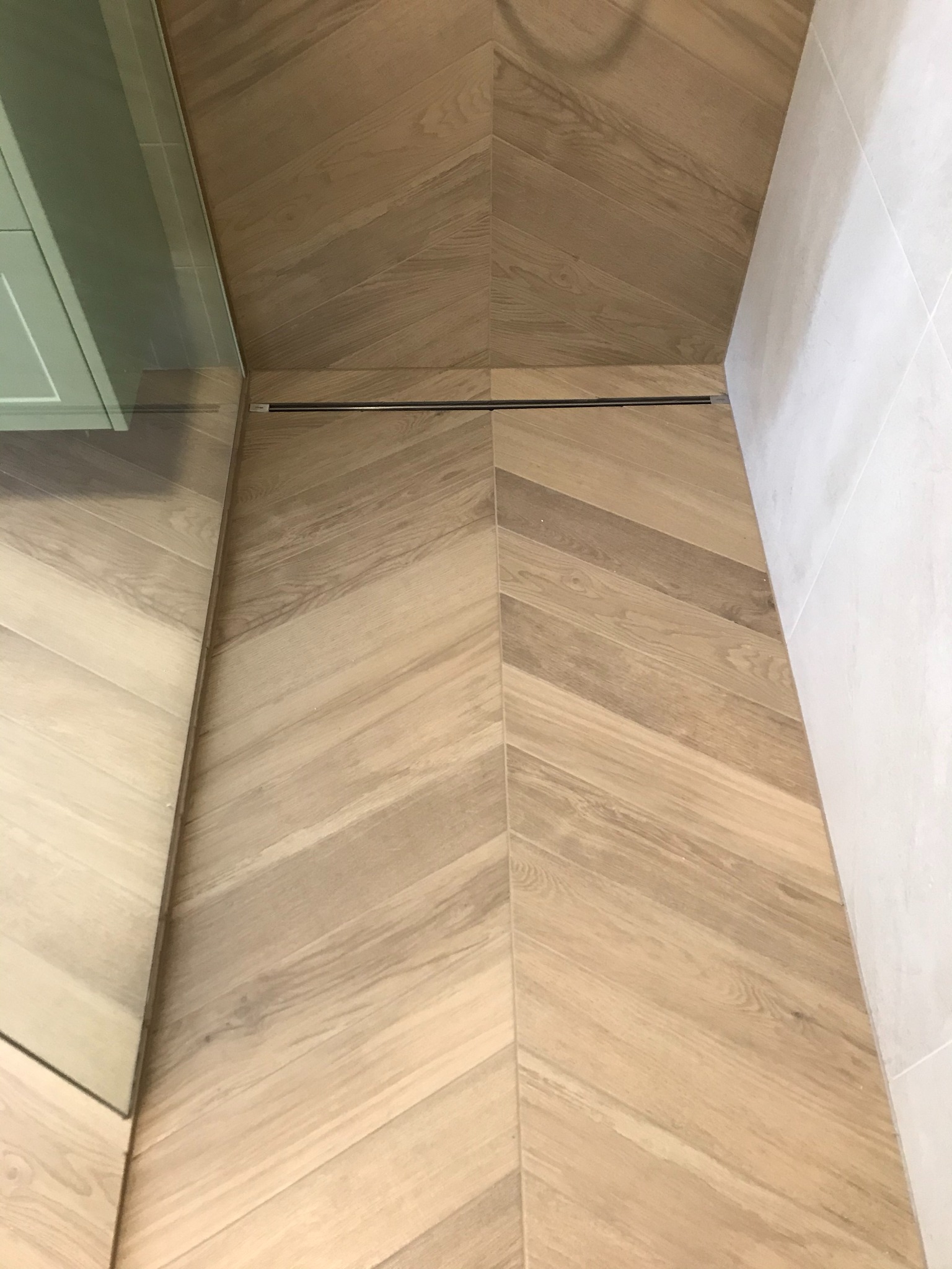 Ułożona w jodełkę, jasna drewnopodobna podłoga w łazience z odpływem liniowym, widok z góry.