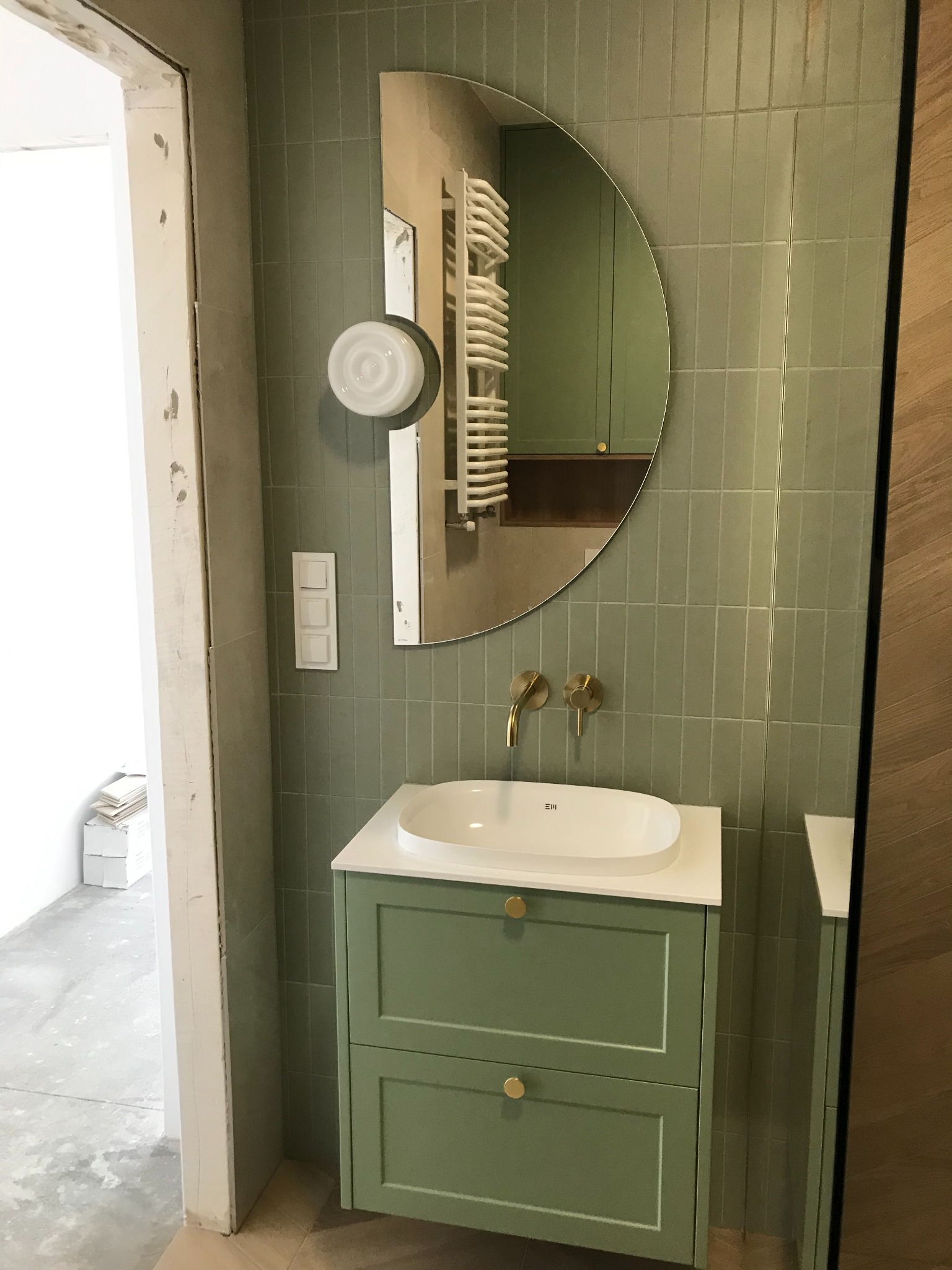 Umywalka nablatowa na szafce w odcieniach zieleni, złota armatura, okrągłe lustro z widocznym grzejnikiem drabinkowym w odbiciu, ściana wyłożona podłużnymi, zielonymi płytkami, widoczne ślady prac...