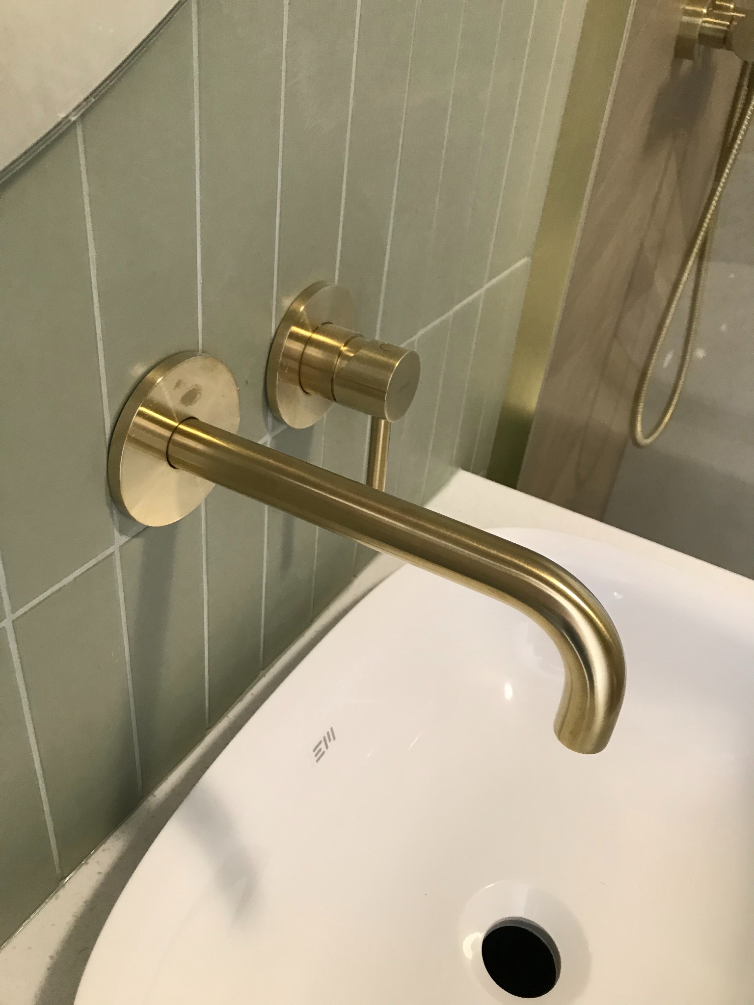 Złota bateria umywalkowa ścienna na tle zielonych płytek w wąskie paski, biała umywalka z widocznym odpływem.