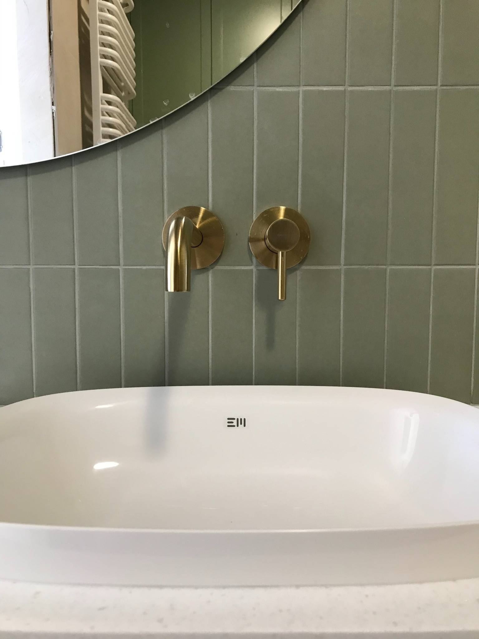 Biała umywalka nablatowa z dwoma złotymi bateriami zamontowanymi na ścianie z zielonych, podłużnych płytek; w tle widoczne lustro owalne i grzejnik drabinkowy.