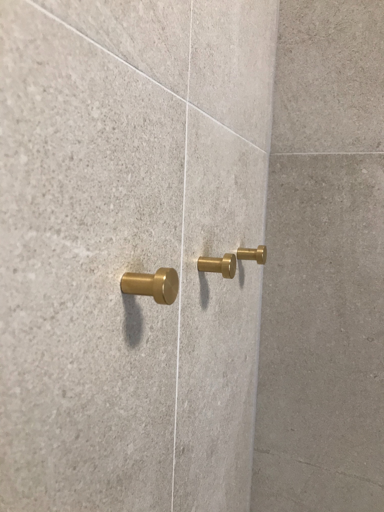 Trzy złote, cylindryczne wieszaki na szarych, kwadratowych płytkach ceramicznych z fugami, ustawione w rzędzie pod kątem.