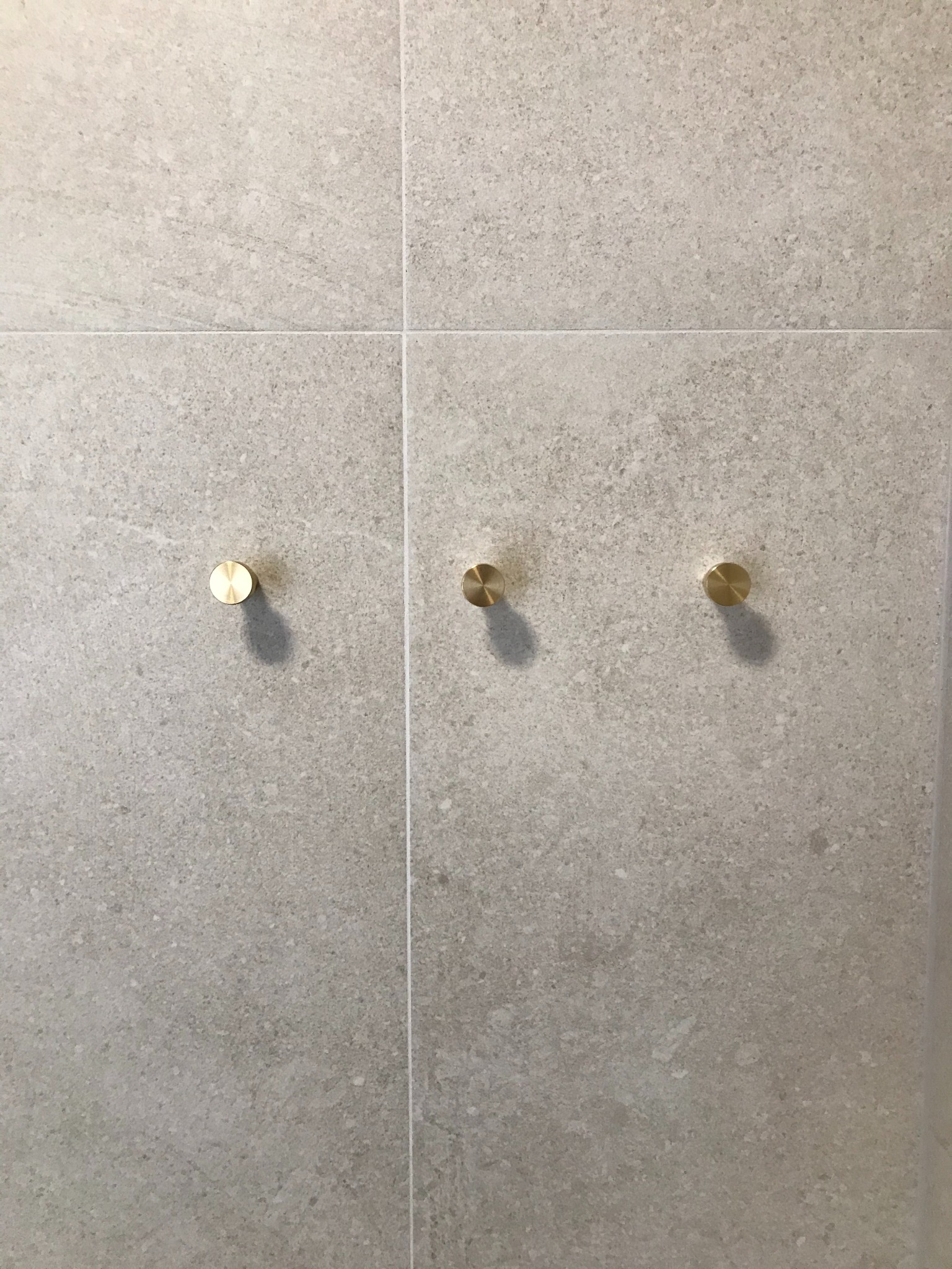 Jasnoszare płytki imitujące kamień z trzema złotymi, okrągłymi wieszakami na ręczniki, zamontowanymi na ścianie.