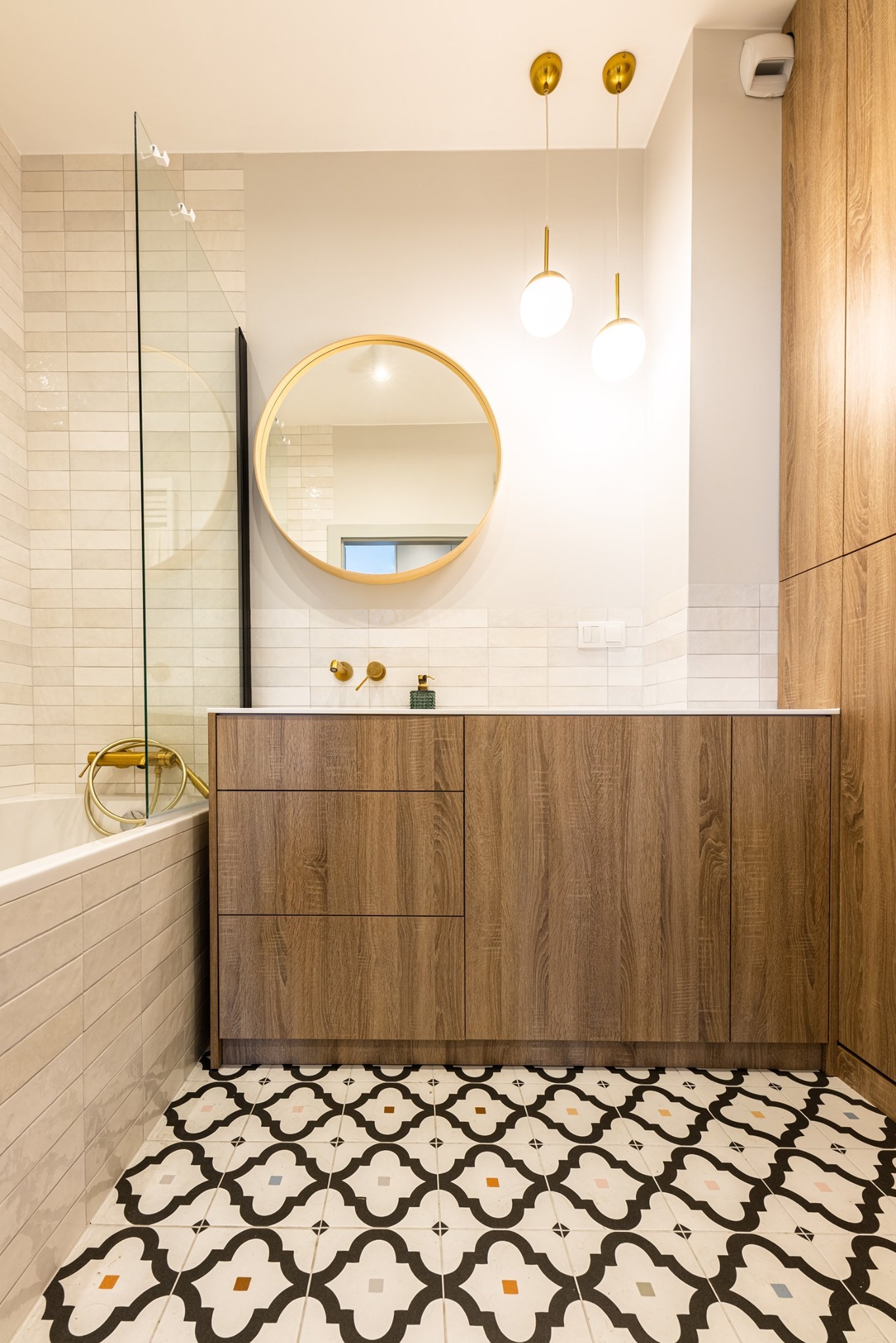 Nowoczesna łazienka z kabiną prysznicową, umywalką z drewnianą szafką, okrągłym lustrem w złotej ramie, złotą armaturą i wzorzystą podłogą.