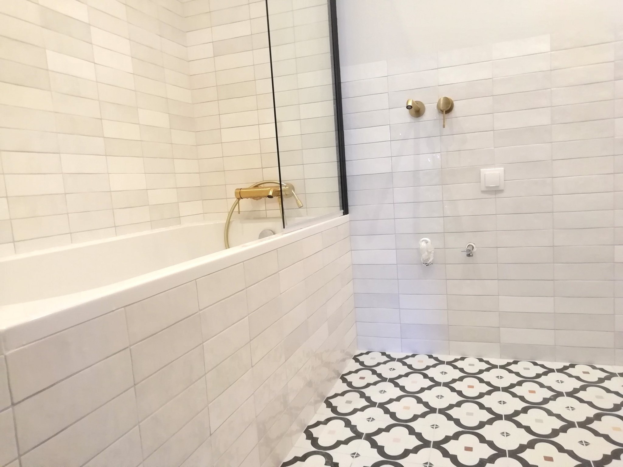 Nowoczesna łazienka z białymi, podłużnymi płytkami na ścianach, złotą armaturą i wzorzystą podłogą w stylu retro.