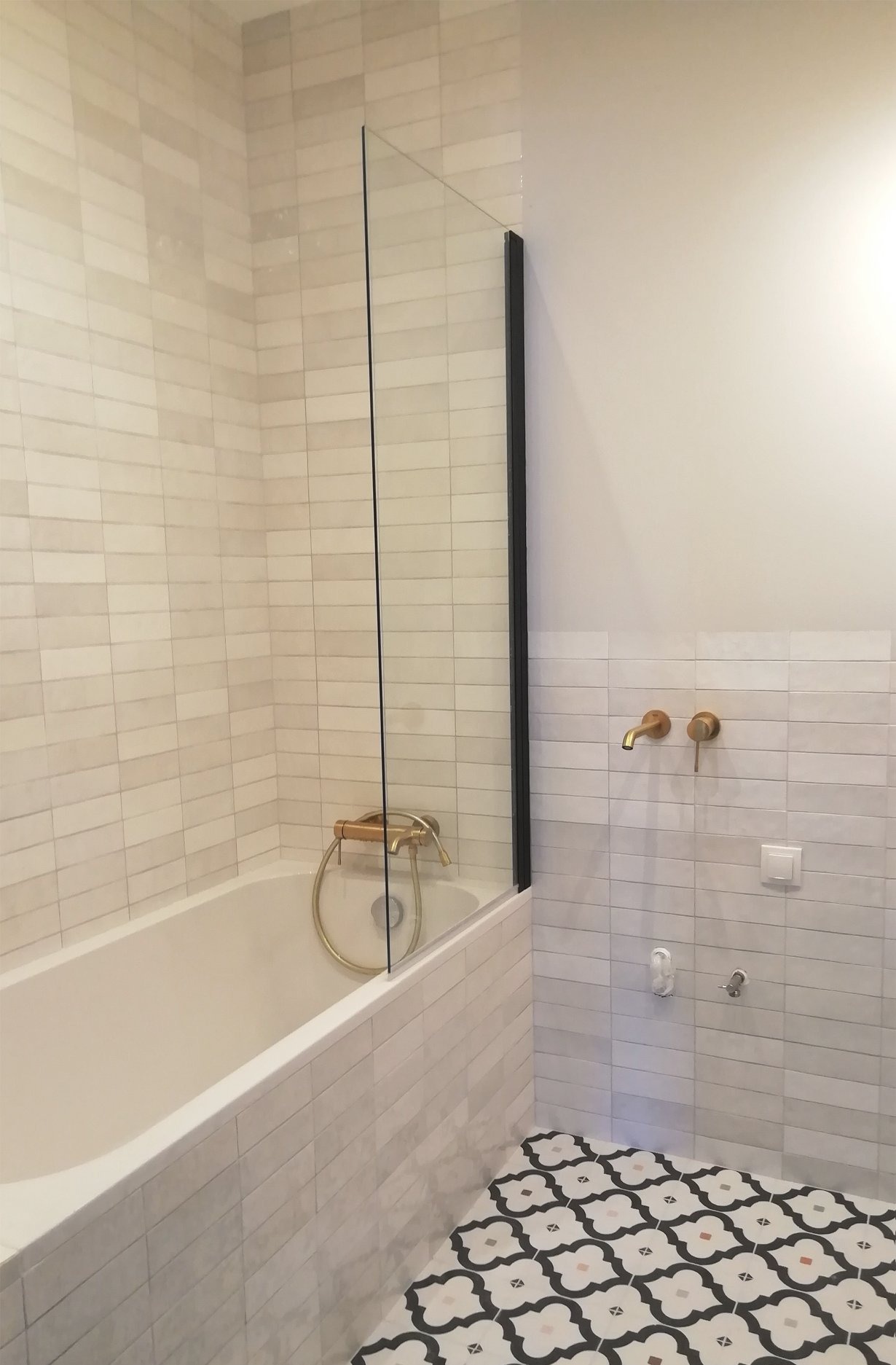 Nowoczesna łazienka w Gdańsku po remoncie, z wanną za szklanym parawanem, złotą armaturą i wzorzystą podłogą.