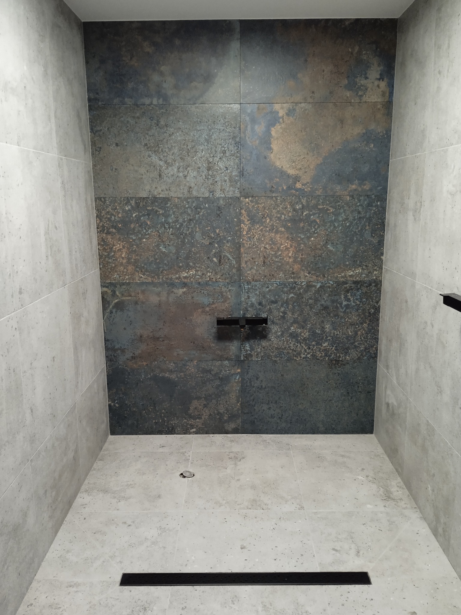 Nowoczesna kabina prysznicowa z płytkami imitującymi rdzę na ścianie i szarymi płytkami na podłodze, widoczny odpływ liniowy i bateria prysznicowa.