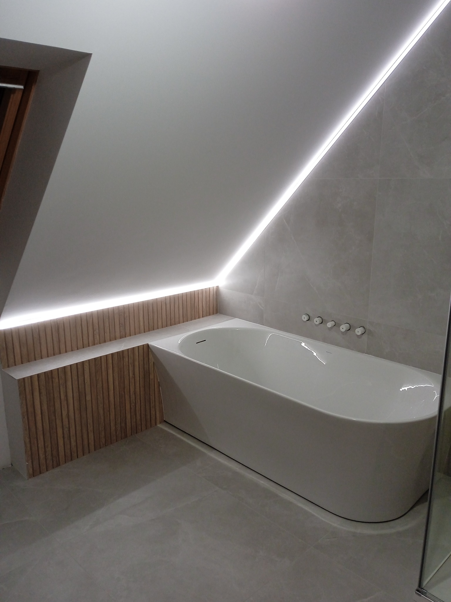 Nowoczesna łazienka na poddaszu z wolnostojącą wanną, podświetleniem LED wzdłuż skosu sufitu i drewnianą zabudową wanny.