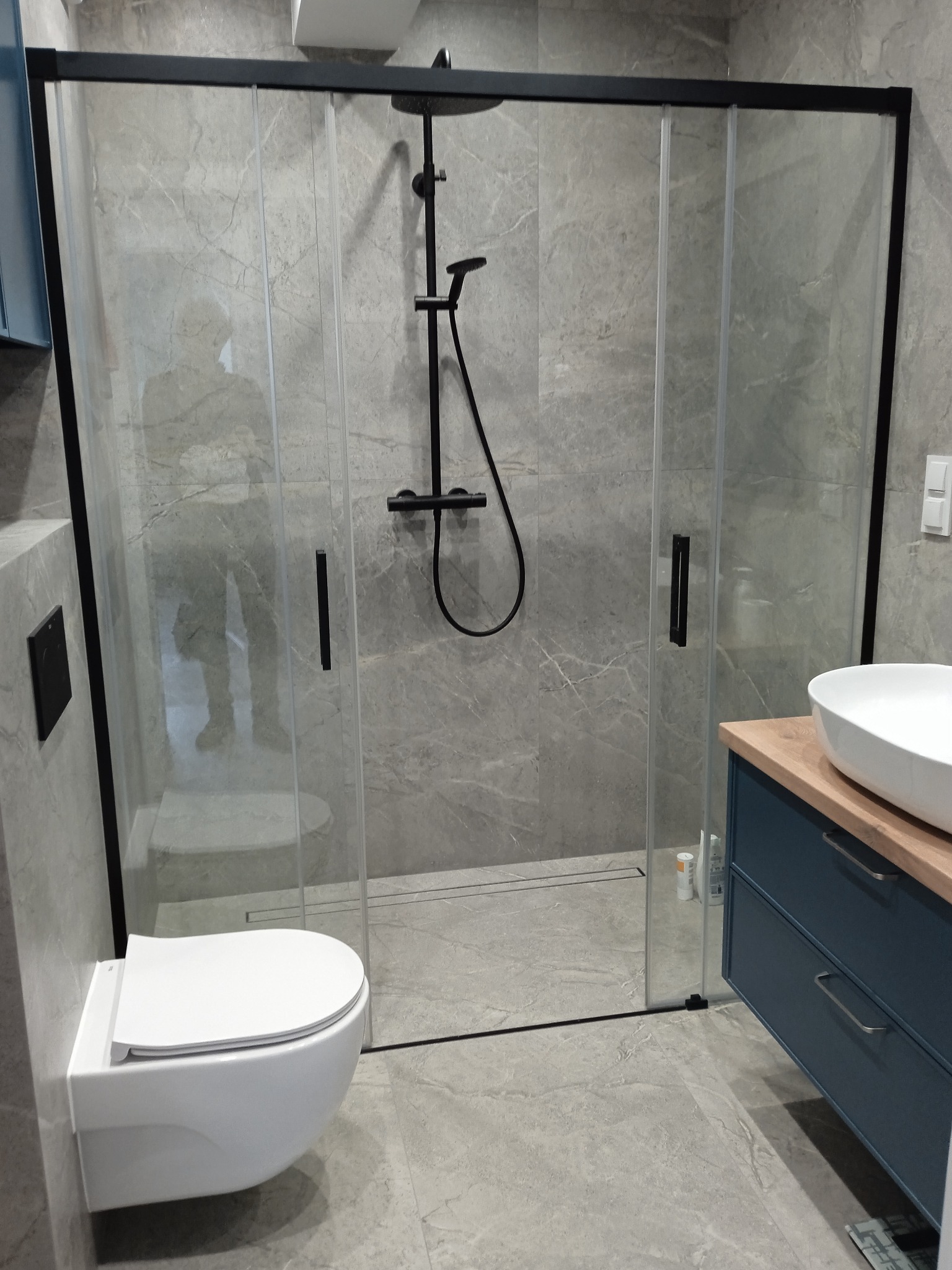 Nowoczesna łazienka z kabiną prysznicową typu walk-in z czarnymi ramami, wiszącą toaletą i szafką umywalkową w kolorze granatowym z drewnianym blatem, wykończona płytkami imitującymi marmur.