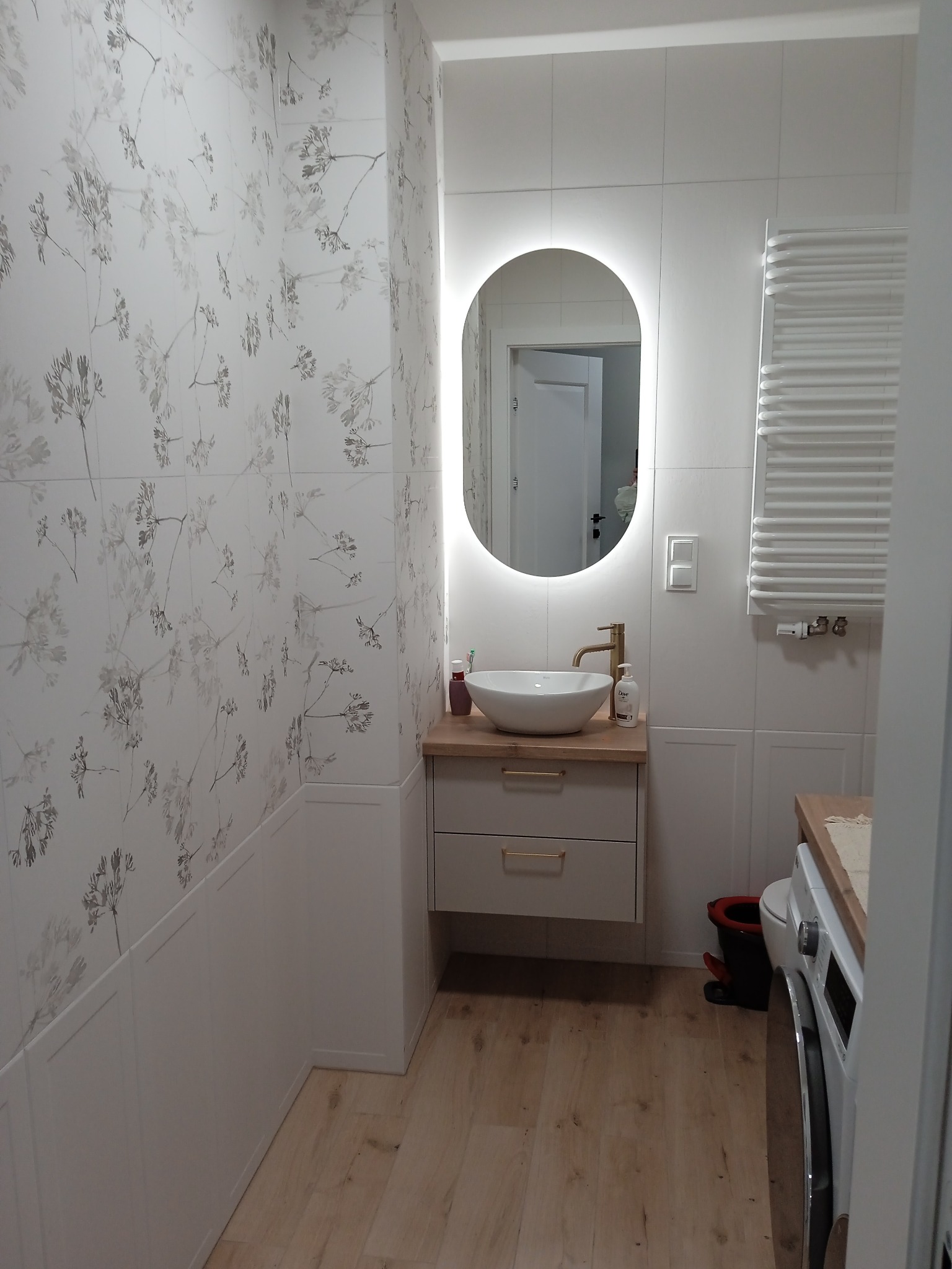 Nowoczesna łazienka z umywalką nablatową, podświetlanym lustrem owalnym i grzejnikiem drabinkowym, wykończona białymi płytkami z motywem roślinnym.