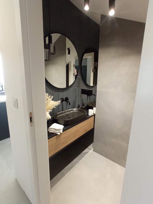 Nowoczesna łazienka w odcieniach szarości i czerni z okrągłymi lustrami, czarną umywalką nablatową i drewnianą szafką, widok zza uchylonych drzwi.