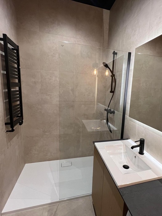 Nowoczesna łazienka z kabiną prysznicową typu walk-in, umywalką nablatową na szafce z drewnopodobnym frontem i grzejnikiem drabinkowym w czarnym kolorze.