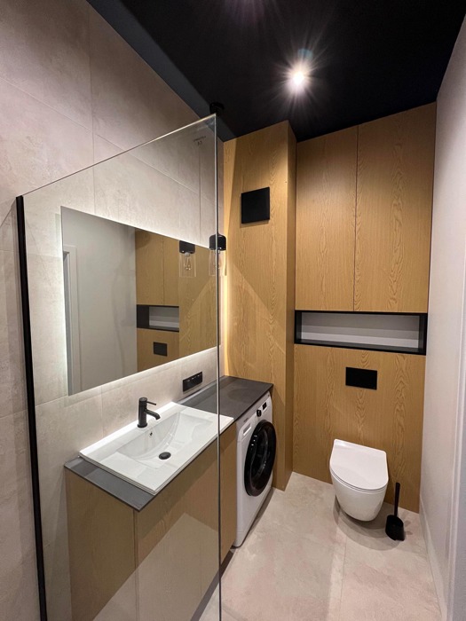 Nowoczesna łazienka z zabudową meblową z jasnego drewna, białą umywalką nablatową z czarną armaturą, pralką obok umywalki oraz toaletą podwieszaną z czarnymi akcentami, widok przez szklaną kabinę...