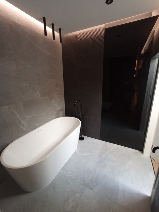 Nowoczesna łazienka z wolnostojącą białą wanną, czarną armaturą i szarą marmurową okładziną ścienną oraz podłogą, oświetlenie LED podsufitowe.