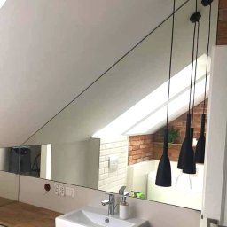 Zakład Szklarski Roman Lizoń - Lustro na skośnej ścianie w łazience z białym zlewem i drewnianym blatem. W odbiciu widoczne lampy wiszące i fragment ceglanej ściany.