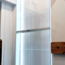 Wysoka, szklana witryna z półkami wkomponowana w białą ścianę. Widoczne delikatne oświetlenie LED wewnątrz. Srebrne okucia i minimalistyczny design.