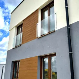 Elewacja dwukondygnacyjnego domu z drewnianą okładziną i szklanymi balustradami balkonowymi, widok z dołu na błękitne niebo z chmurami.