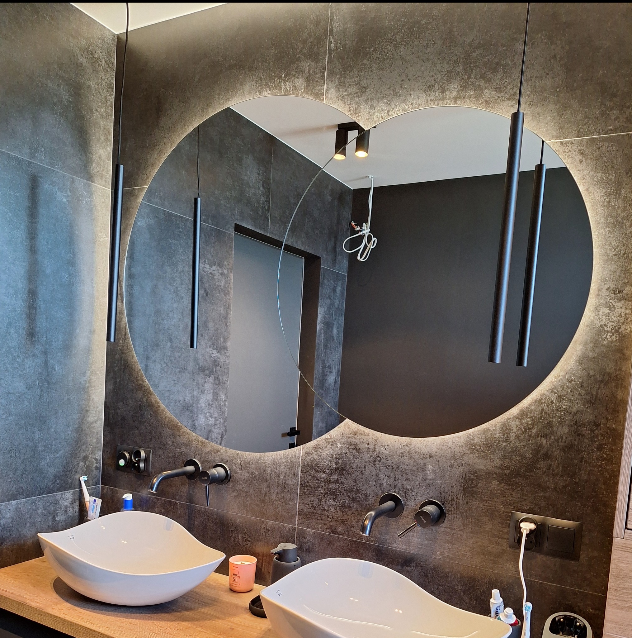 Nowoczesna łazienka z dwoma umywalkami i okrągłym lustrem z podświetleniem LED. Ciemne płytki i minimalistyczne baterie tworzą elegancki wystrój.