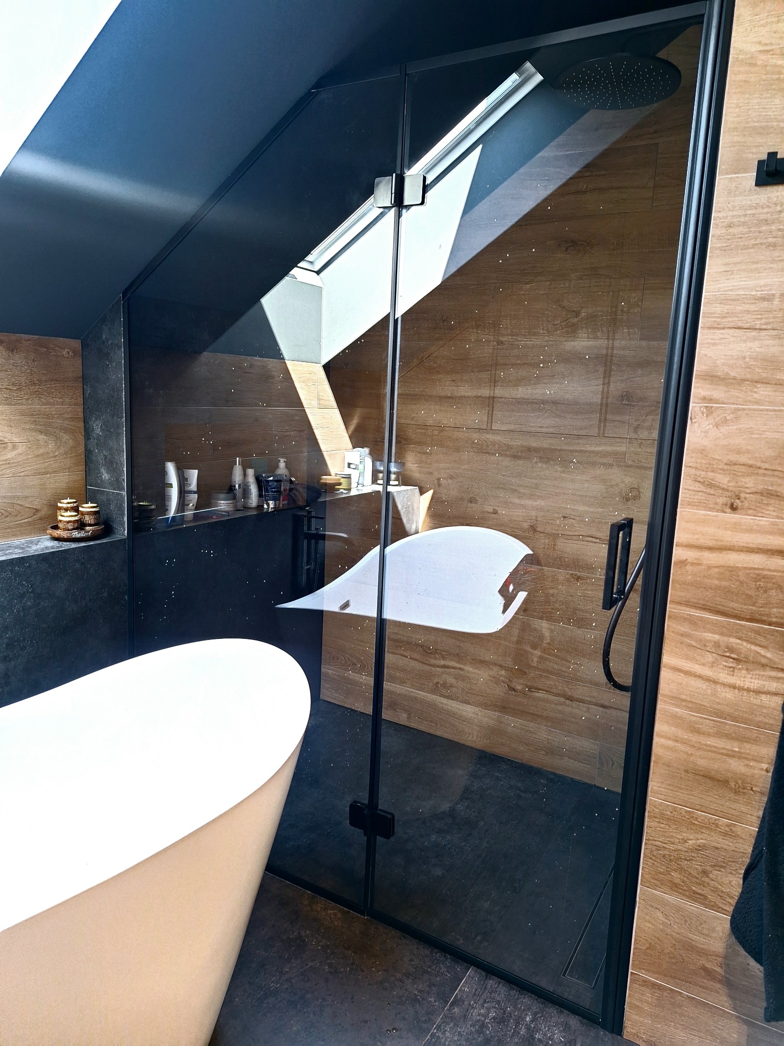 Nowoczesna kabina prysznicowa z czarnymi profilami i dekoracyjną folią na szkle, widoczna obok owalnej wanny w łazience z drewnopodobnymi płytkami.