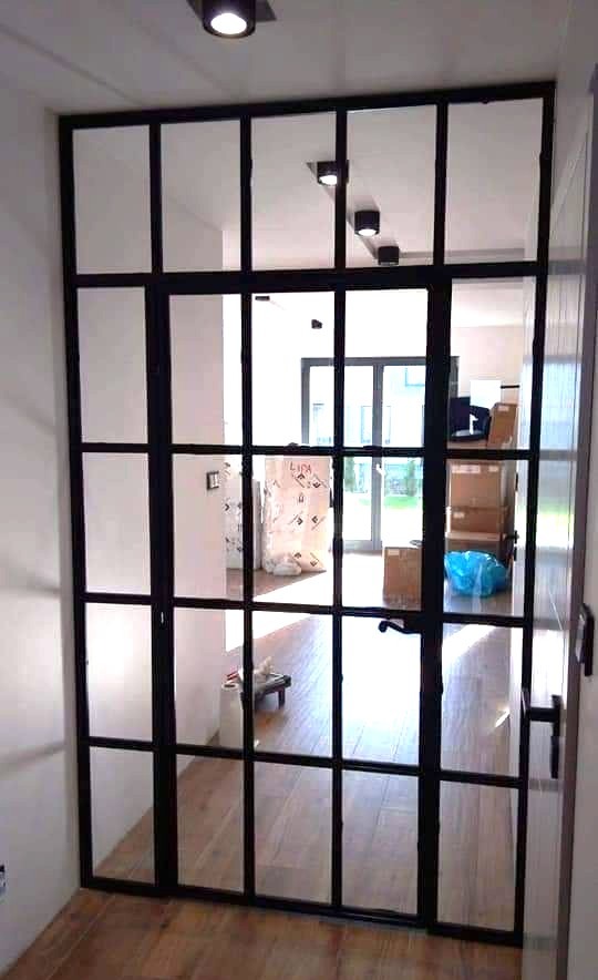 Nowoczesne drzwi wewnętrzne z czarnymi szprosami, dzielące przestrzeń. Widok na jasne wnętrze z drewnianą podłogą i elementami wyposażenia.