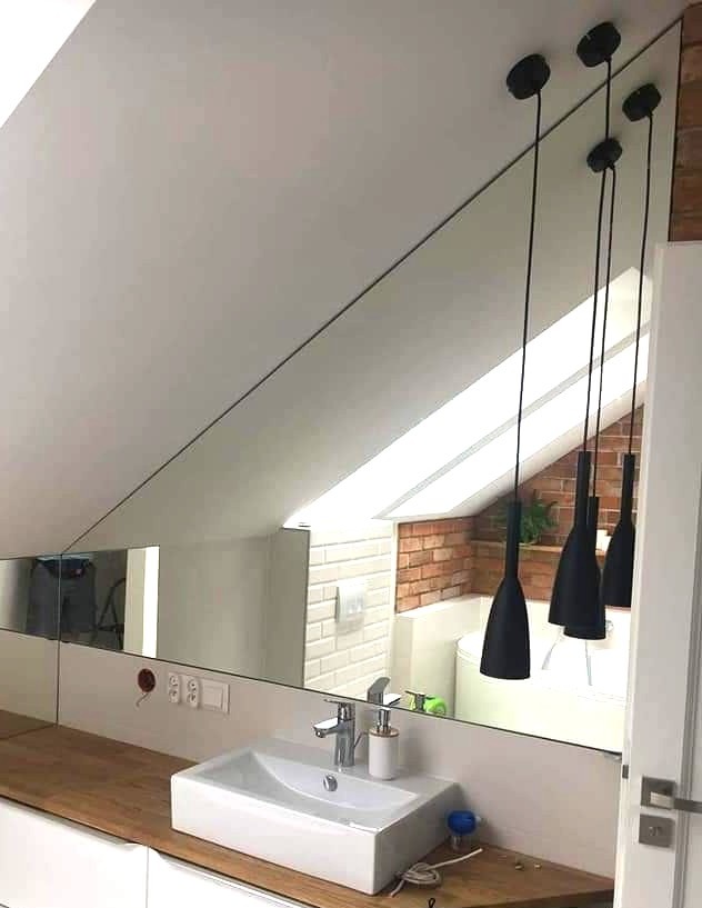 Lustro na skośnej ścianie w łazience z białym zlewem i drewnianym blatem. W odbiciu widoczne lampy wiszące i fragment ceglanej ściany.