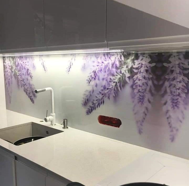 Kuchnia z panelem szklanym nad blatem, zdobionym motywem kwitnącej glicynii. Biała armatura i szafki w tle. Widoczny fragment blatu i zlewozmywak.