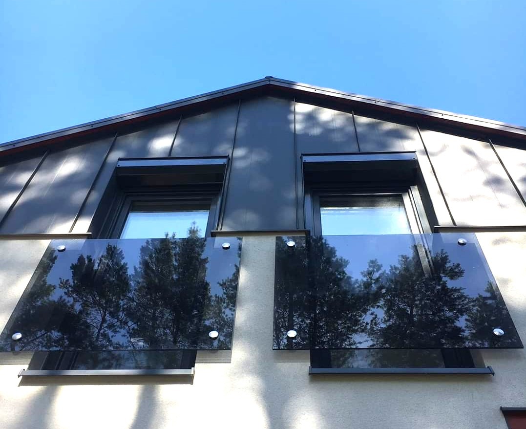 Elewacja budynku z oknami i szklanymi panelami, odbijającymi drzewa. Minimalistyczny design, gra świateł i cieni na fasadzie, błękitne niebo w tle.