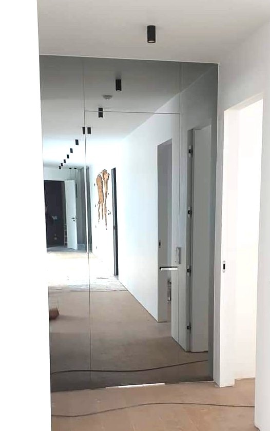 Szklana ściana lustrzana w korytarzu z czarnymi punktowymi lampami sufitowymi odbijająca wnętrze. Widoczne drzwi i fragment instalacji na ścianie.