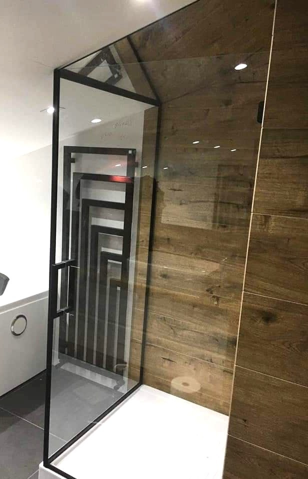 Nowoczesna kabina prysznicowa ze szkła z czarnymi profilami na tle ściany z drewnopodobnych płytek. Widoczny grzejnik drabinkowy i fragment wanny.