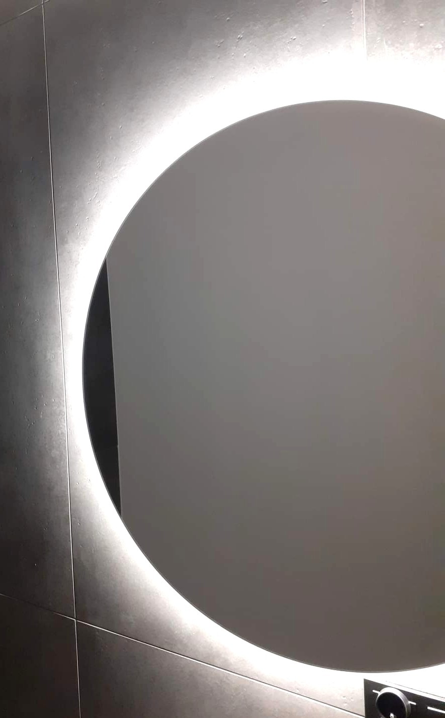 Okrągłe lustro z podświetleniem LED na szarej, płytkowej ścianie. Minimalistyczny design, nowoczesne wykończenie łazienki. Widoczny fragment panelu sterowania oświetleniem.