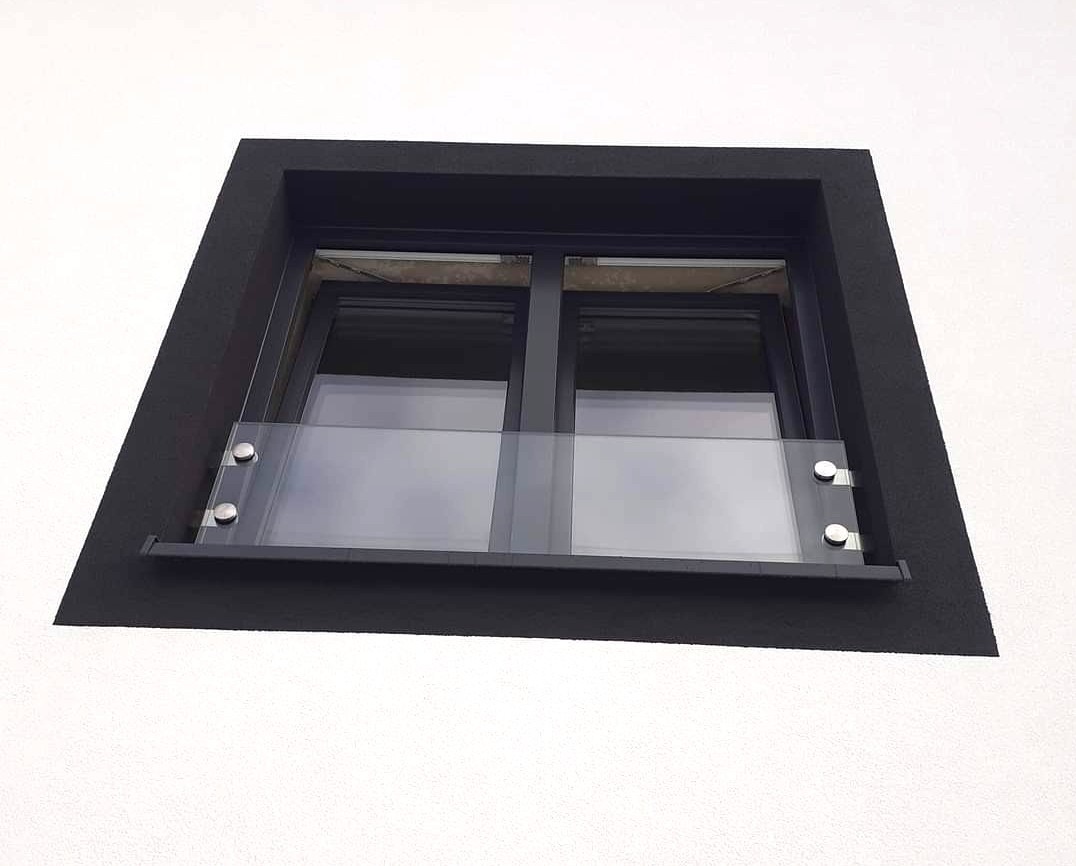 Okno dachowe z czarną ramą i dodatkową szybą ochronną, mocowaną na srebrne uchwyty. Biały, teksturowany mur.