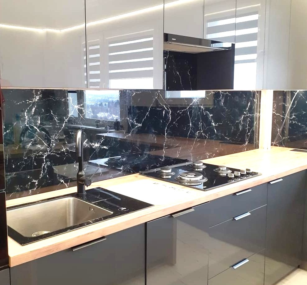 Elegancka kuchnia z czarnym panelem szklanym imitującym marmur nad blatem. Widoczny zlew, płyta grzewcza i błyszczące fronty szafek w odcieniach szarości i bieli.
