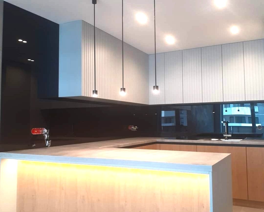Nowoczesna kuchnia z czarnym szklanym panelem nad blatem, oświetleniem LED i wiszącymi lampami. Minimalistyczny design z elementami drewna.