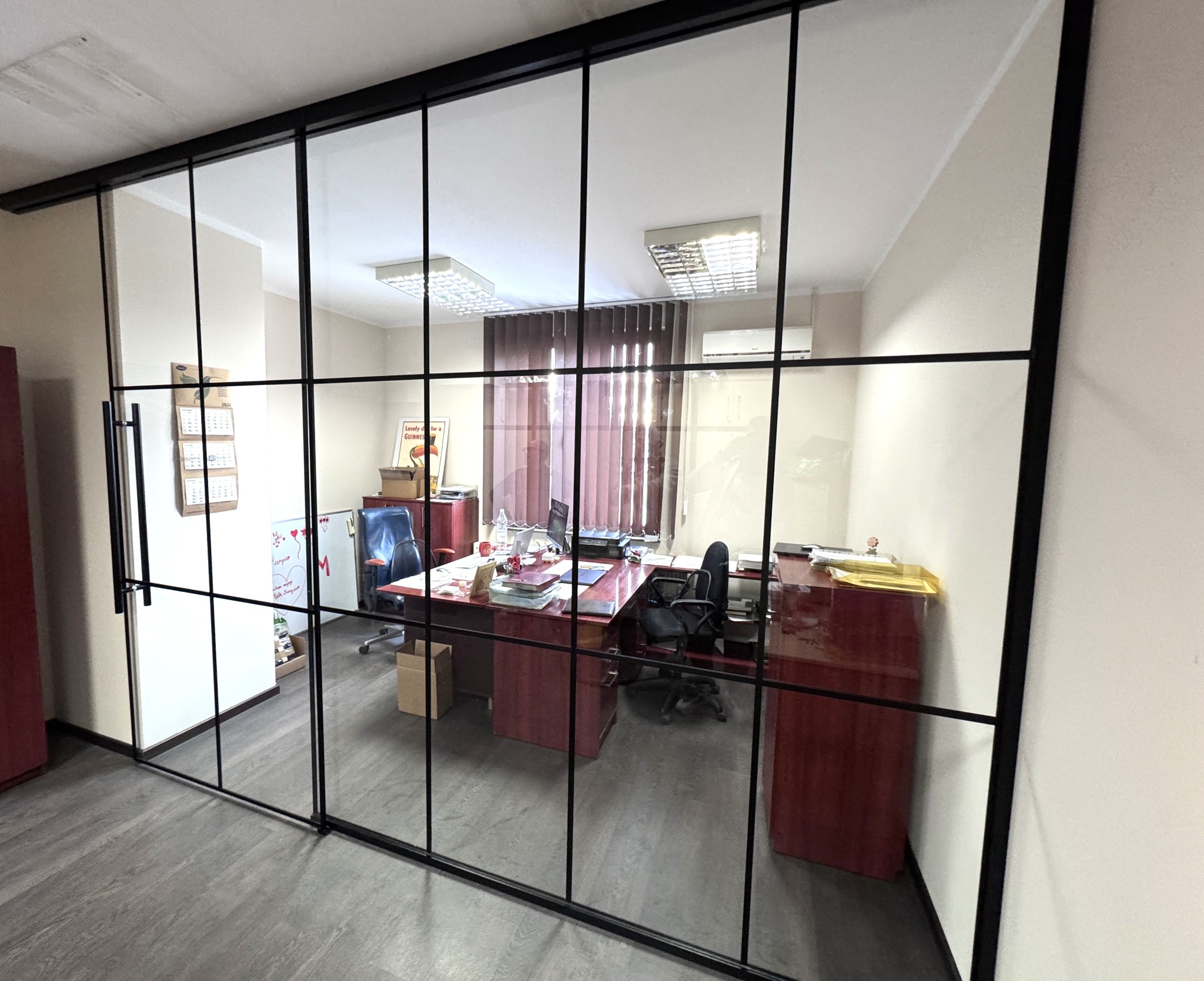 Przeszklona ściana działowa z czarnymi szprosami oddzielająca biuro. Widoczne biurka, krzesła i dokumenty wewnątrz pomieszczenia. Minimalistyczny design.