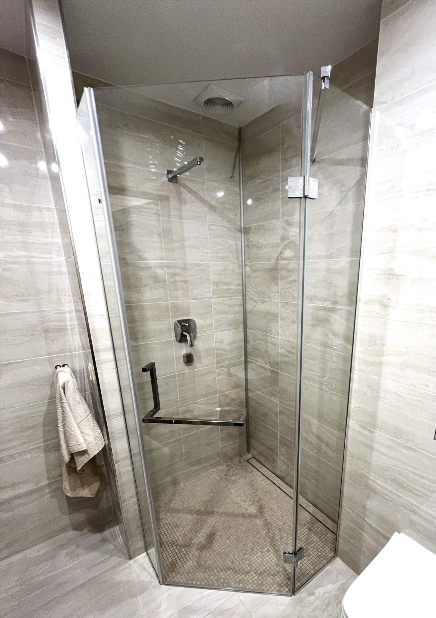 Nowoczesna kabina prysznicowa z przezroczystego szkła, z chromowanymi okuciami, w łazience wykończonej płytkami w odcieniach beżu i szarości. Widoczny odpływ liniowy.