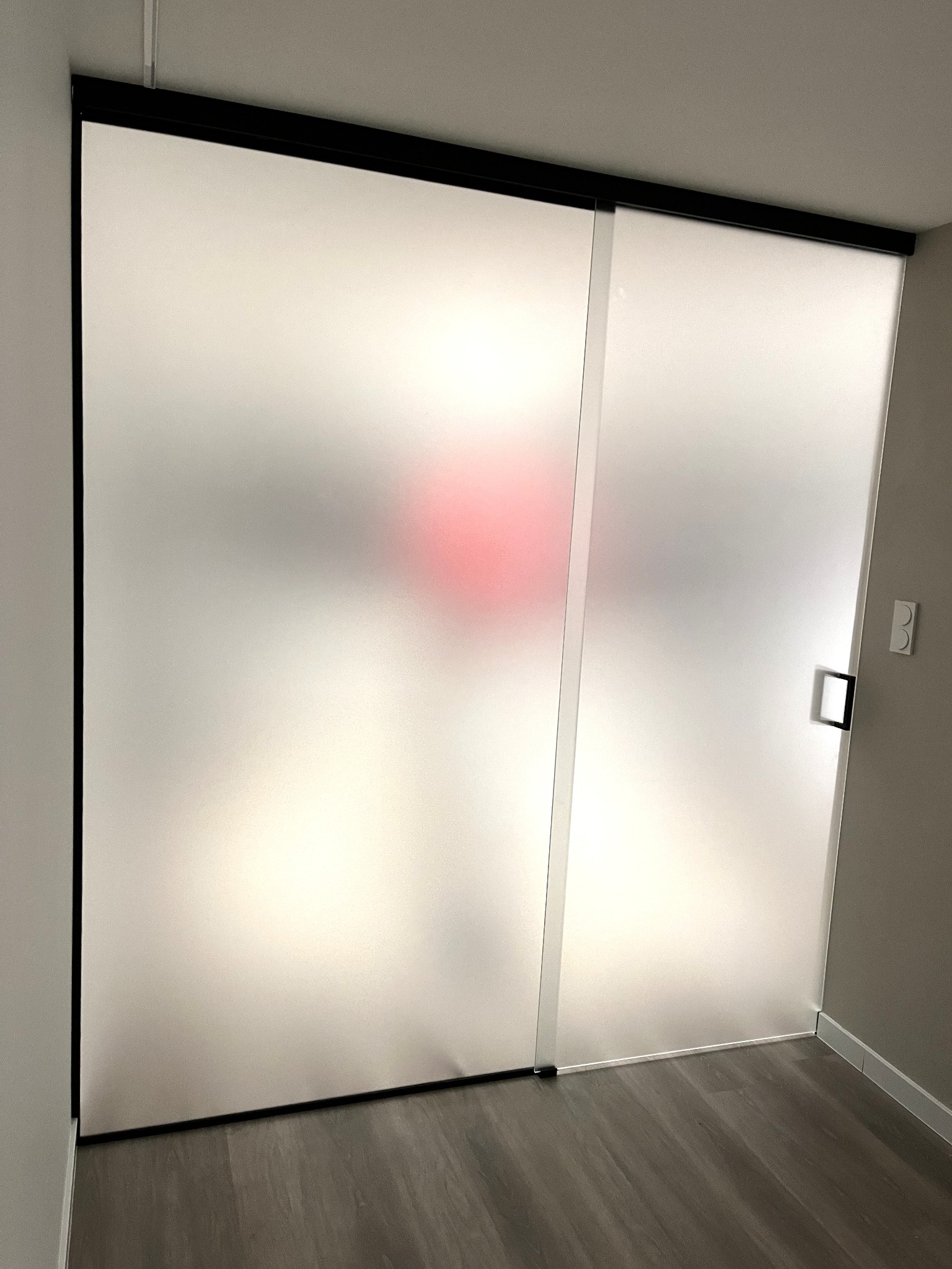 Matowe, przesuwne drzwi z czarną ramą i kwadratowym uchwytem, widoczne na tle jasnego pomieszczenia z drewnianą podłogą. Za drzwiami rozmyty, czerwony kształt.