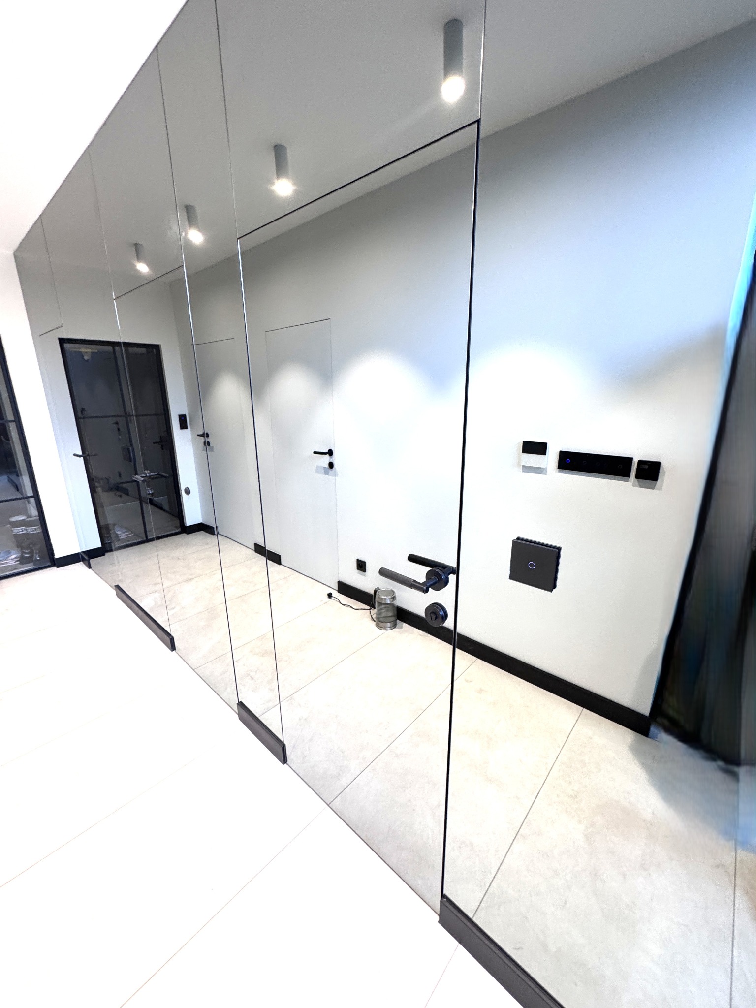 Nowoczesny hol z lustrzanymi ścianami i szklanymi drzwiami, minimalistyczny design, czarne akcenty, subtelne oświetlenie punktowe, jasna podłoga.