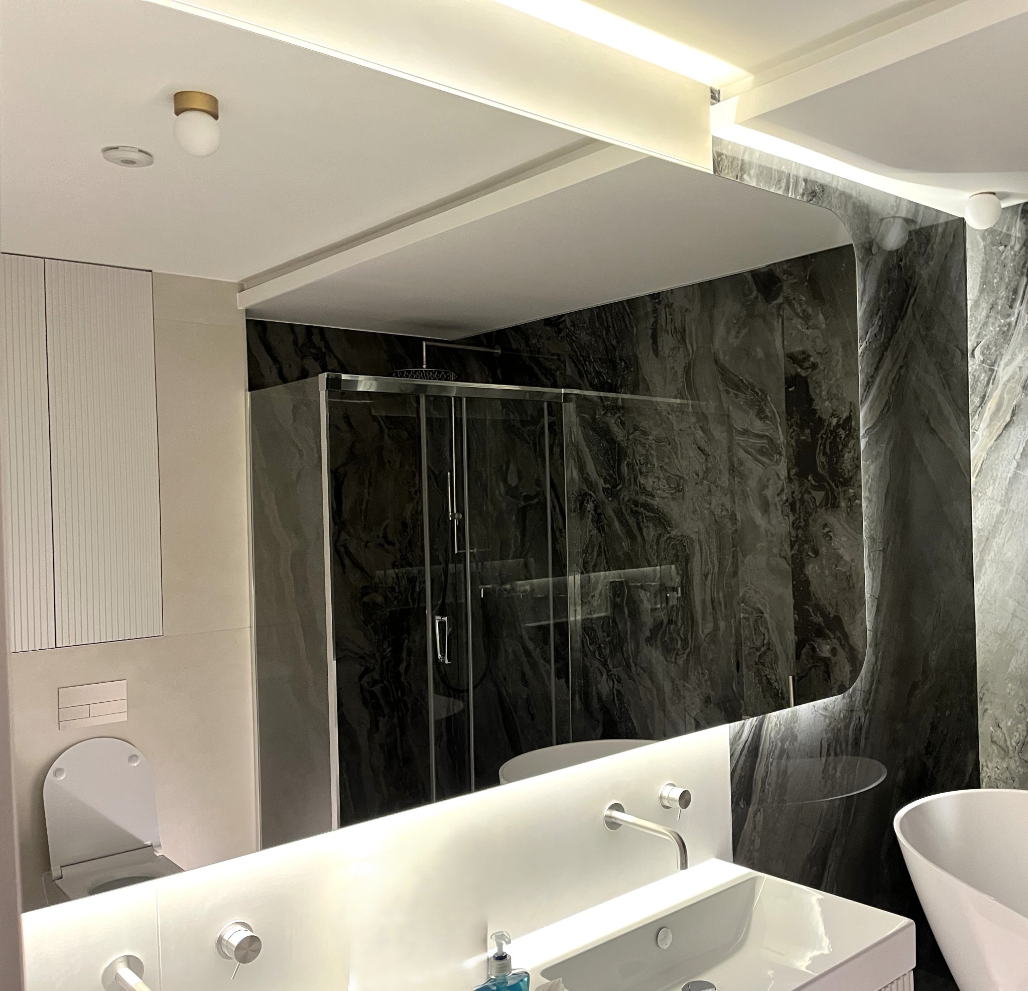 Nowoczesna łazienka z kabiną prysznicową za szklaną ścianą, marmurową ścianą i umywalką z podświetlanym lustrem. Minimalistyczny design i eleganckie wykończenie.